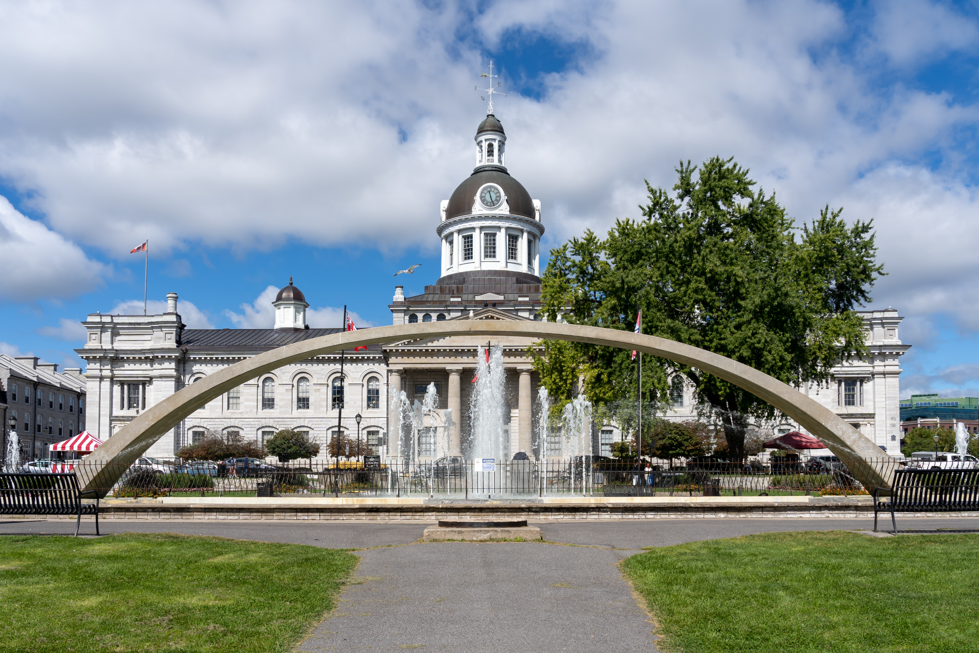 A large limestone building with a prominent central cupola and dome containing a clock and weather vane. In front, a large, contemporary arched concrete water fountain. 